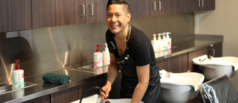 Federico student looking and smiling at the camera as he washes a client's hair.